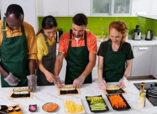 What a New Orleans Hands-on Cooking Class Looks Like Four diverse adults wearing aprons participate in a sushi-making class in a modern kitchen.