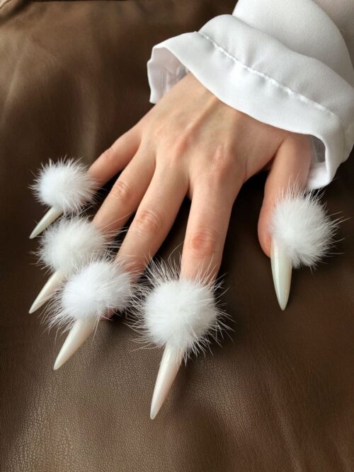 A hand with long, pointed white acrylic nails decorated with fluffy white pom-poms at the base, resting on a brown leather surface.