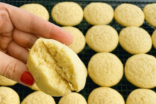 Hand holding a soft vanilla cookie with a bite taken, above a tray of similar cookies.