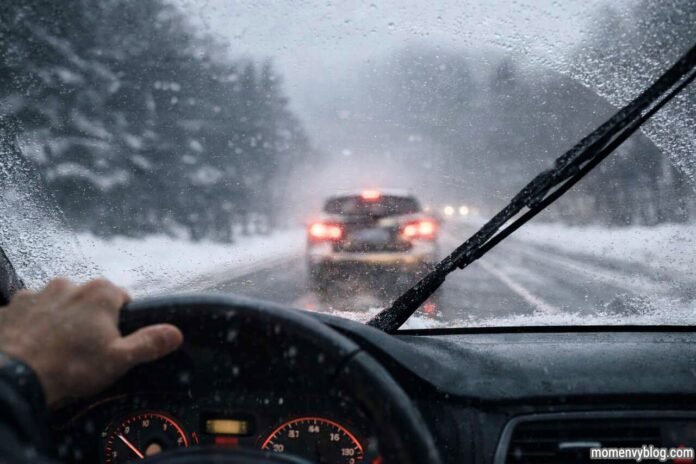 Driver’s view through a car windshield during heavy snow and rain, with windshield wipers clearing water and a vehicle ahead with red brake lights on a slippery road.
