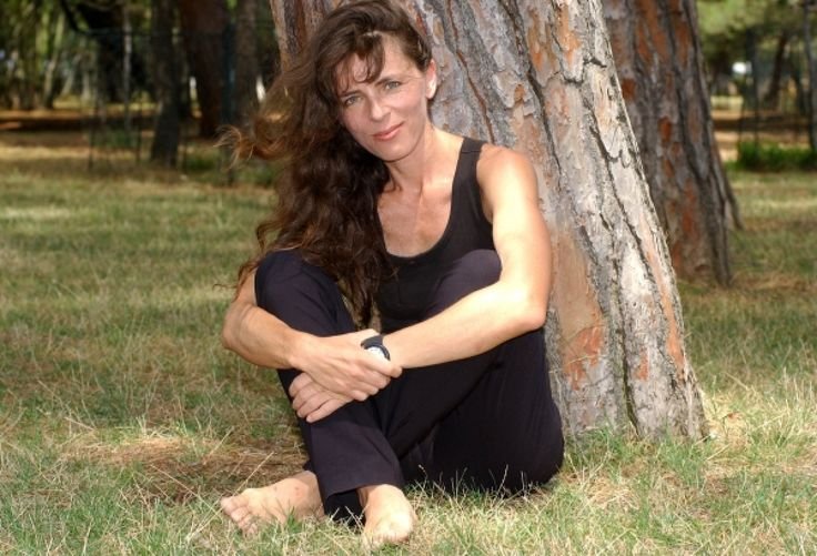 Mira Furlan with long dark hair sitting barefoot on grass and leaning against a large tree in a park, wearing a black sleeveless outfit and looking toward the camera.