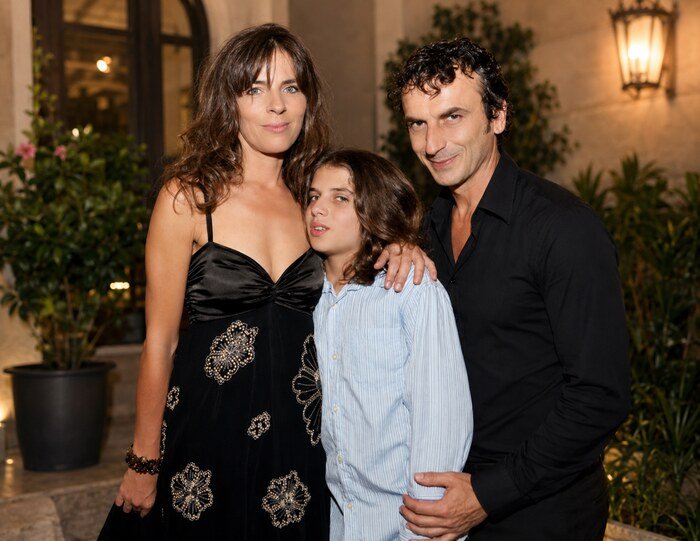 Family portrait of marko lav gajić and Mira Furlan with a child posing together outdoors in the evening, standing in front of a warmly lit building entrance with plants in the background.